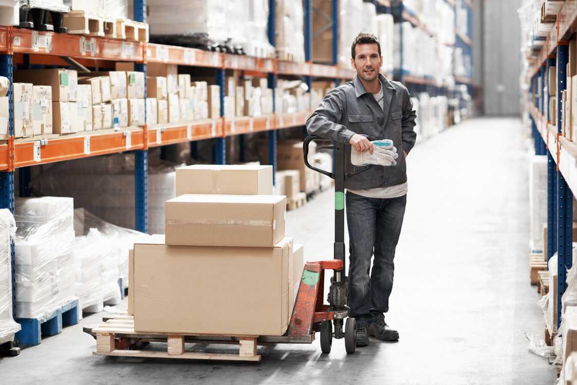 A warehouse operator with a loaded cart beside high racking in a distribution aisle